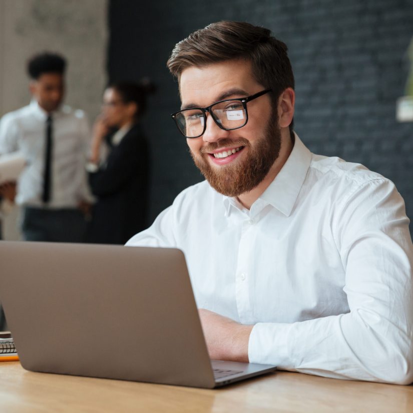 Photo of happy young caucasian businessman sitting indoors using laptop computer. Looking camera.